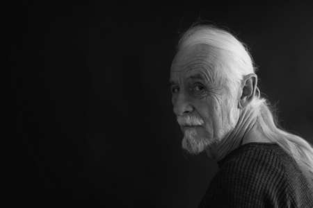 Low key studio black and white portrait of beautiful old man with gray hair in a braid looking over his shoulder at the camera. Horizontally.の写真素材