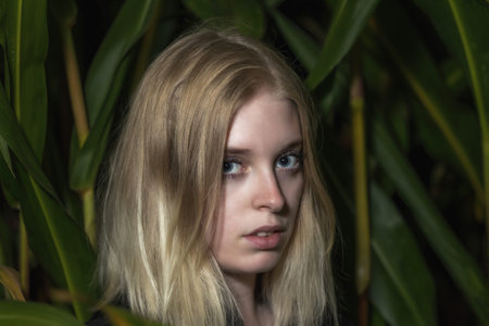 Blonde young woman poses among the green plants. Horizontally.