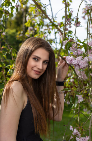 Attractive brunette girl dressed in a black dress is holding a sprig of cherry blossom. Vertically.の写真素材