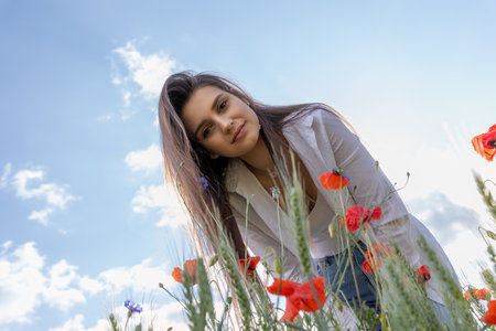 Smiling brunette girl is looking down at the camera in a field of red poppies. The blue sky is in the  background.. Horizontally.の写真素材
