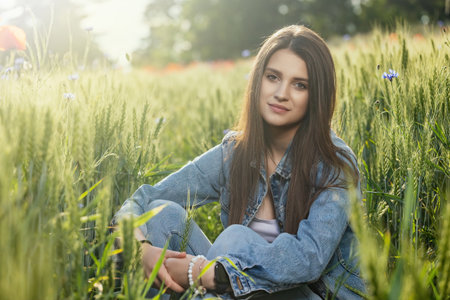 Beautiful brunette girl dressed in jeans is sitting in green cornfield looking at the camera. Horizontally.の写真素材
