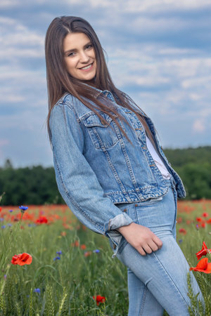 Smiling long haired  brunette girl dressed in blue jeans is posing in red poppies meadow. Vertically.の写真素材