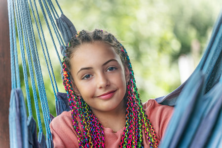 Smiling young girl with colorful braids in her hair is posing on blue hammock. Horizontally.の写真素材