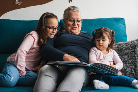 A grandmother with her two Caucasian granddaughters, on the sofa in her house, reading a fairy tale to them. The three of them are very happy and smiling.の写真素材