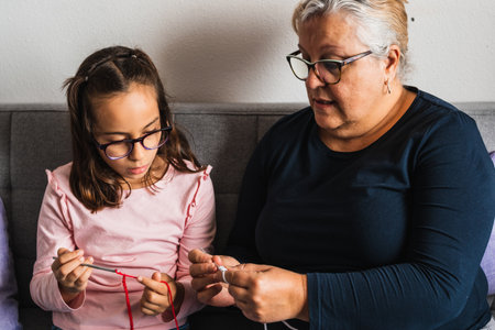 Grandmother teaching her granddaughter to knit with needle, both sitting on the sofa in the living room at home.の写真素材
