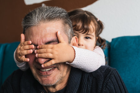Granddaughter covers her grandfather's eyes from behind, without him seeing her, to surprise him, they both smile happily.の写真素材