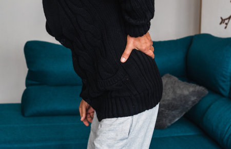An elderly man touches his back after getting up from the sofa, due to severe pain located in the...