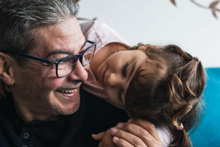 Granddaughter hugs her grandfather from behind, from behind. They are both on the sofa at home. And they smile very happy to be together.の写真素材
