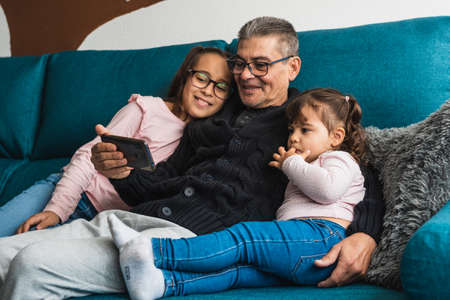 A grandfather with his two granddaughters, sitting on the sofa at home, while they look at the mobile smiling.の写真素材