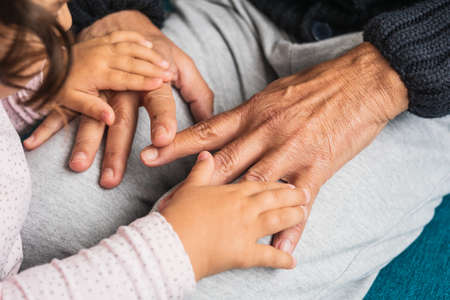 Close-up of the wrinkled hands of a grandfather grasping the little hands of his granddaughter with love and delicacy.の写真素材
