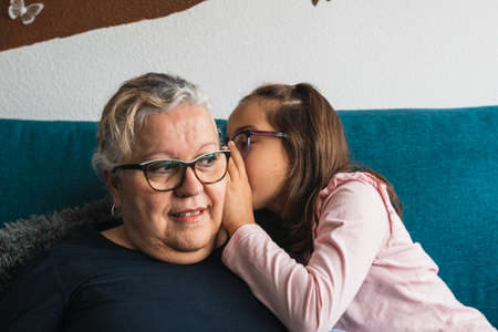 Granddaughter who tells a secret to her grandmother, who listens carefully. Both of Caucasian race. They are on the sofa in the living room at home.の写真素材