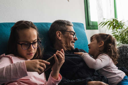 A grandfather with his two granddaughters, sitting on the sofa at home, while they look at the mobile smiling.の写真素材