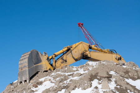 The arm and bucket of a hydraulic excavator on top of a pile of earth in winter with a crane in the...