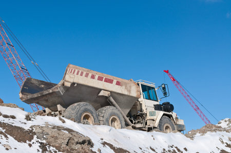 A dump truck sits on top of a pile of earth at a construction site in winter with two cranes in the...
