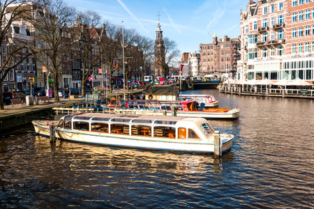 Editorial stock picture of tourist ferries in one of the canals in Amsterdam. Three empty boats lie side by side. It is a beautiful day in early April, and the trees still stand without leaves. Great marketing picture for Amsterdam in the spring.のeditorial素材