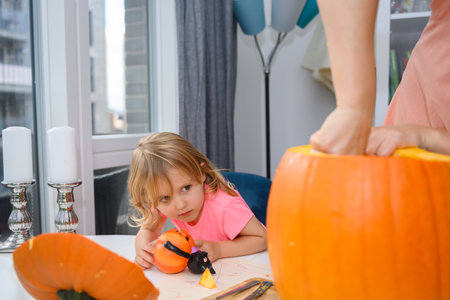Cute little Caucasian girl helping her mother with cutting out a pumpkin for Halloween. They are sitting inside, cutting out the pumpkin af their dinner table.の写真素材