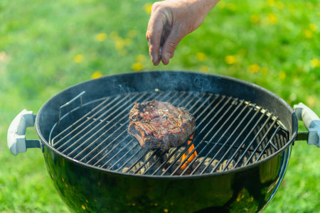 Delicious, juicy sirloin sizzling on a black grill, in spring sunlight, with lush green lawn with dandelions in the background. A person is pouring coarse salt on the steakの写真素材
