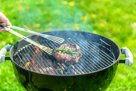 Delicious, juicy sirloin sizzling on a black grill, with a lot of fire and smoke in the grill. There is a lush green lawn with dandelions in the background. A hand with a grill tong reaches in to grab the steak. There is rosemary on the steakの写真素材
