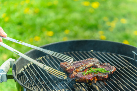 Delicious, juicy sirloin roasting on a traditional black grill, in spring sunlight, with lush green lawn with dandelions in the background. A hand reaches in to grab the steak with a grill tong. There is rosemary on the steakの写真素材