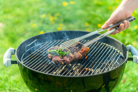 Delicious, juicy steak roasting on a traditional black grill, in spring sunlight, with lush green lawn with dandelions in the background. A hand reaches in the place the steak with a grill tong. There is rosemary on the steakの写真素材