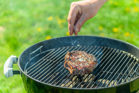 Delicious, juicy sirloin sizzling on a black grill, in spring sunlight, with lush green lawn with dandelions in the background. A person is pouring coarse salt over the sirloinの写真素材