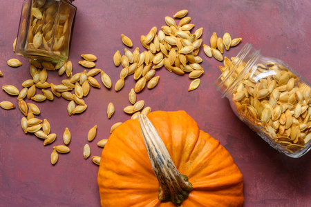 Two glass jars with roasted pumpkin seeds lying on a rustic table. A cut off top of a pumpkin is lying besides.の写真素材