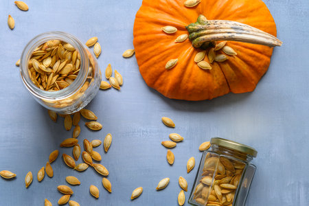 Two glass jars with roasted pumpkin seeds lying on a rustic table. A cut off top of a pumpkin is lying besides.の写真素材