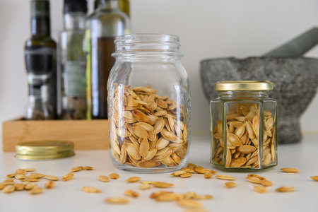 Two glass jars with roasted pumpkin seeds on a kitchen table. Oil and vinegar bottles and a mortar and pestle in the backgroundの写真素材