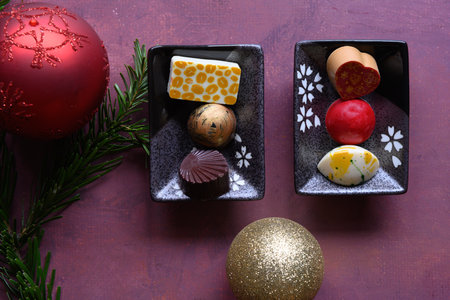 Two small plates with 3 pieces of delicious homemade chocolate on each, on a rustic burgundy background with copy space around. Christmas balls and spruce green on the left side of the platesの写真素材
