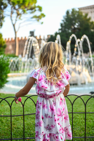 Small, cute girl with curly golden hair, in a beautiful white dress with pink flowers, admiring a small fountain in a park in Verona, Italy. The girl has her back to the camera. It is a beautiful sunny summer dayの写真素材