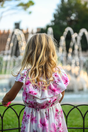 Small, cute girl with curly golden hair, in a beautiful white dress with pink flowers, admiring a small fountain in a park in Verona, Italy. The girl has her back to the camera. It is a beautiful sunny summer dayの写真素材