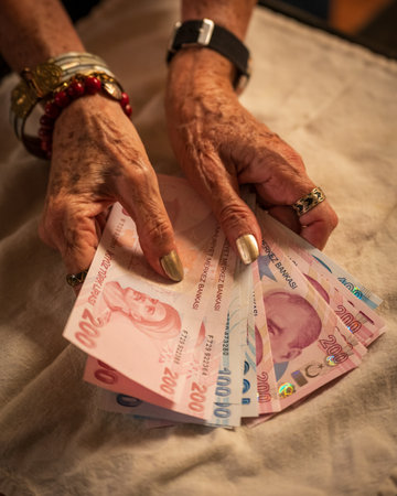 An elderly woman holding several Turkish lira banknotes, including 200 and 100 lira bills. Their hands are adorned with bracelets and rings, and they appear to be counting or organizing the money.の写真素材