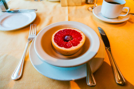 A simple breakfast plate featuring a halved grapefruit topped with a single blueberry. The table is set with silverware, coffee, and plates on a soft yellow tablecloth.の写真素材