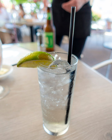 A tall glass of gin and tonic, garnished with a fresh lime wedge, sits chilled on a cafÃ© table. The crisp drink sparkles in focus, with the background fading in soft, dreamy blur.の写真素材