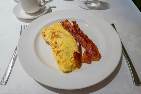 Breakfast plate featuring a fluffy omelette alongside crispy strips of bacon, elegantly presented on a white tablecloth with cutlery and a coffee cup in the background, perfect for a morning meal.の写真素材