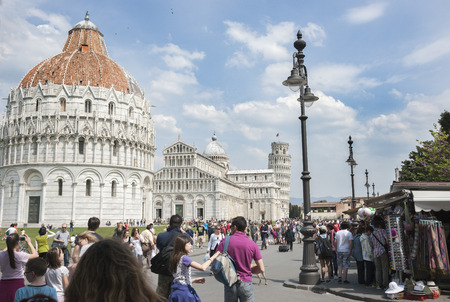Pisa, Italy - April 23, 2011: Tourists moving along the Piazza del Duomo in sight of the Baptistry, the Duomo Del Pisa and the Leaning Tower and past the souvenir stalls on right in Pisa, Italy.のeditorial素材
