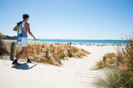 MOUNT MAUNGANUI, NEW ZEALAND-JANUARY 22 2015; youth walks to beach on hot summer day, carrying his...