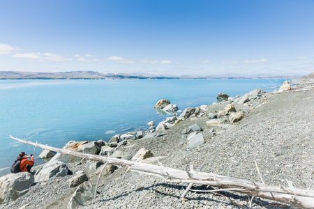 Canterbury, New Zealand; February 15, 2015; Lake Pukaki, Canterbury, South Island New Zealand. The turquoise water, blue sky and grey stony foreshore with a tourist sitting amongst rocks taking photograph.のeditorial素材