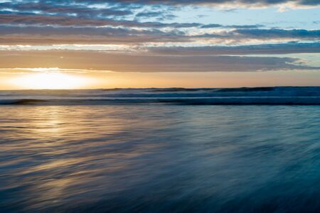 Waitarere  Beach Levin New Zealand the distant surf and wash of sea on beach smoothed by long exposure in blue tones and golden setting sunの写真素材