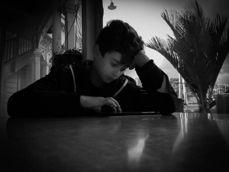 Grainy effect black and white boy at table in low light in front of window resting  on his elbow concentrating with hand poised over his mobile deviceの写真素材
