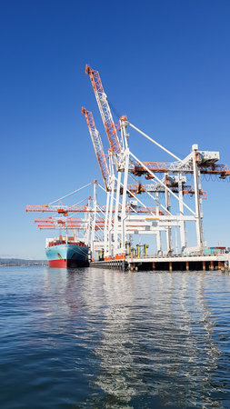 Tauranga, New Zealand - April 19, 2017;  Port of Tauranga container terminal with tall white and orange cranes stretching up and out over moored ship and reflected in deep blue harbour water.のeditorial素材