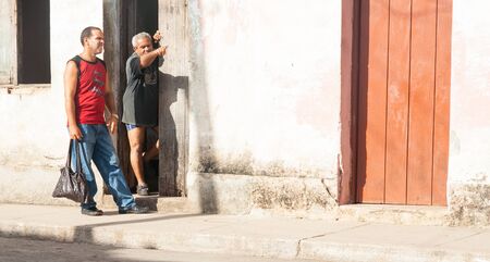 Trinidad de Cuba - July 2, 2012 ; Man standing in doorway of Spanish style home in Cuba points way to another in red tank top standing on footpath in Cuban town.のeditorial素材