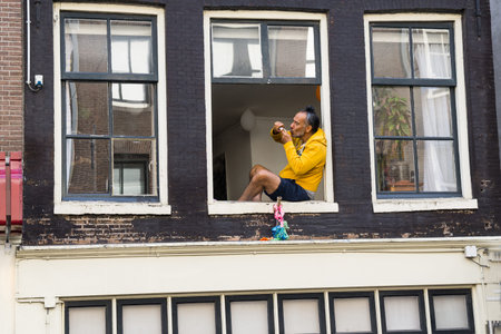 AMSTERDAM, HOLLAND - AUGUST 21, 2017; Man in yeloow hoodie top sititing in window on first floor apartment lights up.のeditorial素材