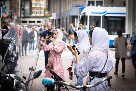 AMSTERDAM, HOLLAND - AUGUST 21, 2017; Three young Muslim women in white hijab in busy city street standing pre-occupied using mobile phonesのeditorial素材