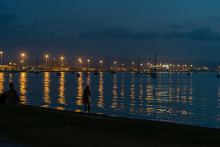 Silhouette of people on beach with port lights reflected between leisure boats moored on calm water of bay.の写真素材