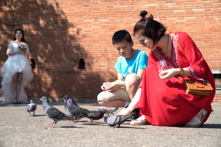 CHIANG MAI,  THAILAND - JANUARY 28 2018;  Woman in bright red dress crouches feeding pigeons in Pratu Tha phae Park outside the old city wall east gate with bride in background shadows being photographed..のeditorial素材