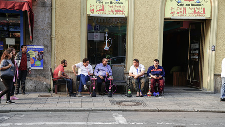 MUNICH GERMANY -SEPTEMBER 8, 2017; Middle Eastern men in Munich side street sitting outside shop on footpath talking and smoking or vaporising flavored tobacco in hookah pipes as people walk by.のeditorial素材
