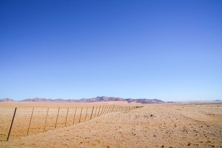 Namibia wide open desert landscape divided by long fence heading towards distant hills with red sand drifts.の写真素材