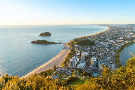 Mount Maunganui stretches out below as sun rises on horizon and falls across ocean beach and buildings belowの写真素材