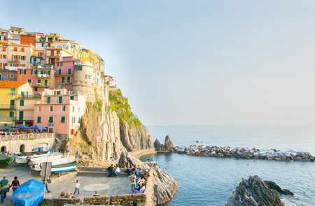MANAROLA ITALY - APRIL 25 2011; Boats  pulled up and people on square below steep colored hillside village buildings built on rock cliff face in Cinque Terre on Mediterranean seaのeditorial素材